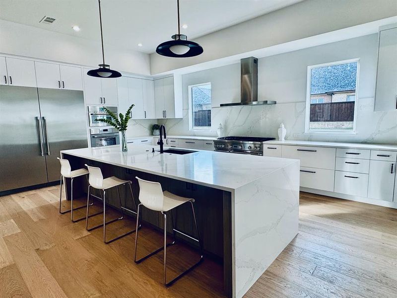 Kitchen with appliances with stainless steel finishes, backsplash, wall chimney range hood, a center island with sink, and hanging light fixtures