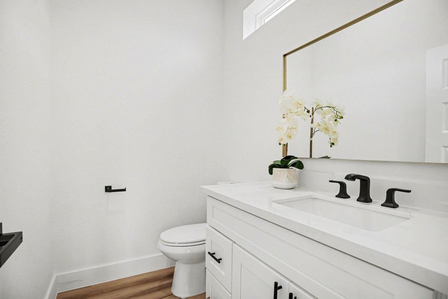 This photo showcases a modern and bright half bathroom with white walls and wood flooring. It features a sleek white vanity with black fixtures, a large mirror, and a potted plant for decoration.