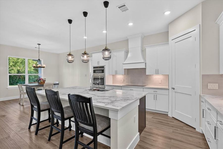 Open-concept kitchen featuring an oversized island with countertop seating, stainless steel appliances including a built-in oven and microwave, custom range hood, and wood-finish flooring Open-concept kitchen featuring an oversized island with countertop seating, stainless steel appliances including a built-in oven and microwave, custom range hood, and wood-finish flooring