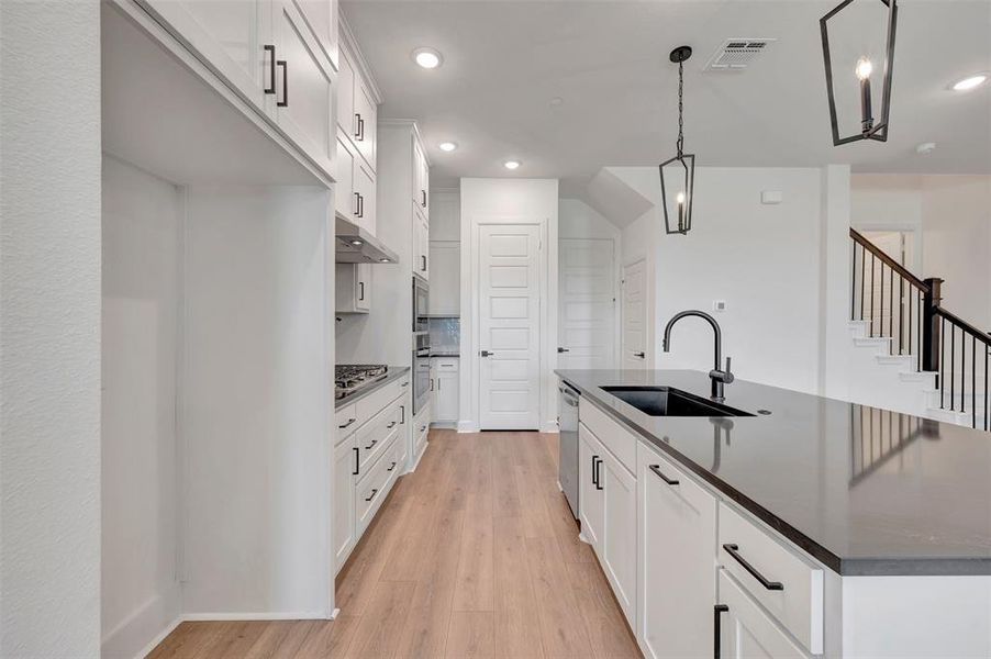 Spacious kitchen featuring white cabinetry with matte black hardware, wood-finish flooring, a large island with an undermount sink and dark countertop, stainless steel appliances, and recessed lighting Spacious kitchen featuring white cabinetry with matte black hardware, wood-finish flooring, a large island with an undermount sink and dark countertop, stainless steel appliances, and recessed lighting