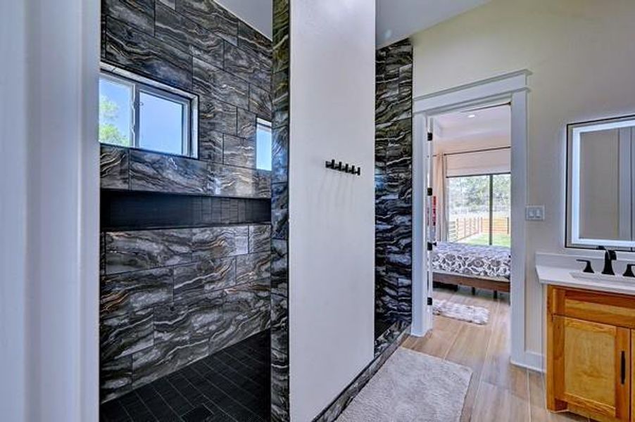 This photo showcases a modern bathroom with a striking dark stone-tiled shower, dual windows for natural light, and a wooden vanity. The space connects to a brightly lit bedroom with hardwood flooring, enhancing the home's contemporary feel.