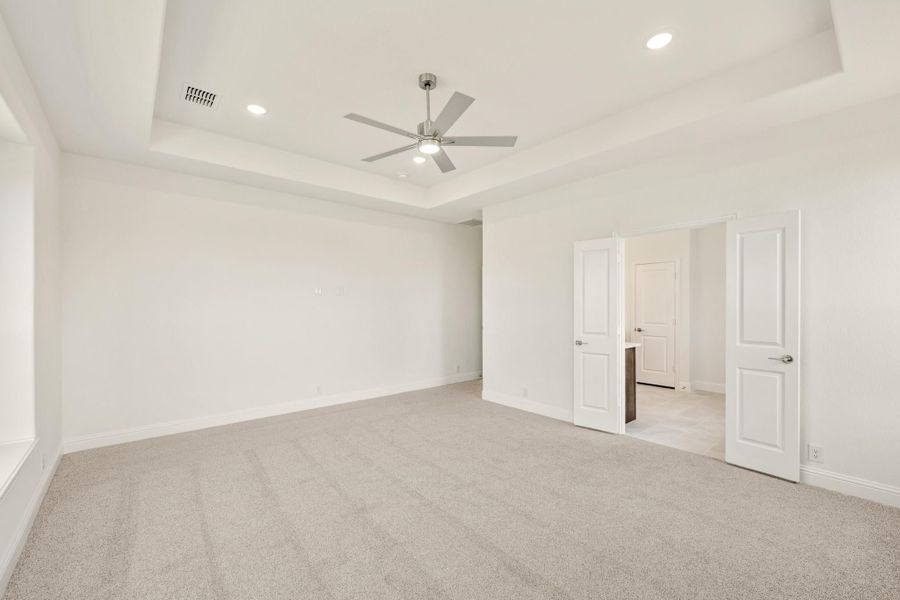 Empty bedroom with tray ceiling, ceiling fan, carpet flooring, and open doors to adjoining rooms Empty bedroom with tray ceiling, ceiling fan, carpet flooring, and open doors to adjoining rooms
