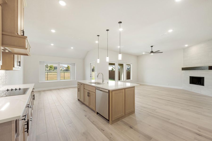 Open kitchen with center island, pendant lights, light wood floors, and adjacent living area with fireplace