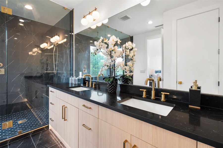 Bathroom with dual vanity featuring light wood cabinetry, black countertops, and gold-toned fixtures