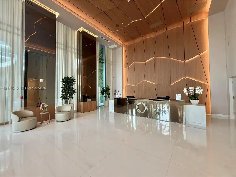 Double-height lobby featuring polished stone flooring, intricate wood-paneled accent wall with integrated ambient lighting, floor-to-ceiling sheer window treatments, and metallic reception desk