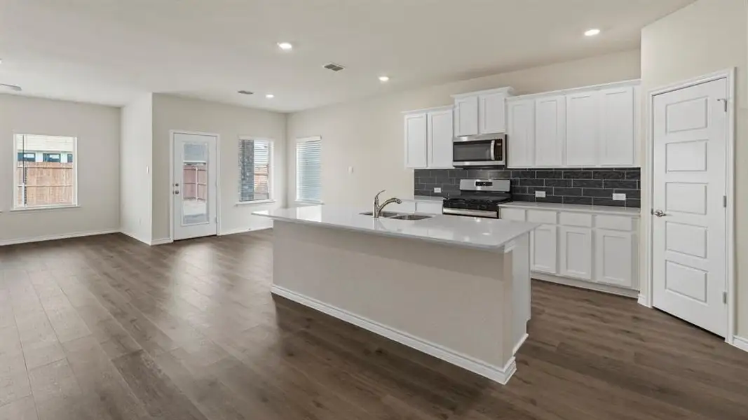 Open-concept living space featuring wood-finish flooring, a kitchen island with an integrated sink, white cabinetry, a stainless steel microwave and range, and a dark subway tile backsplash