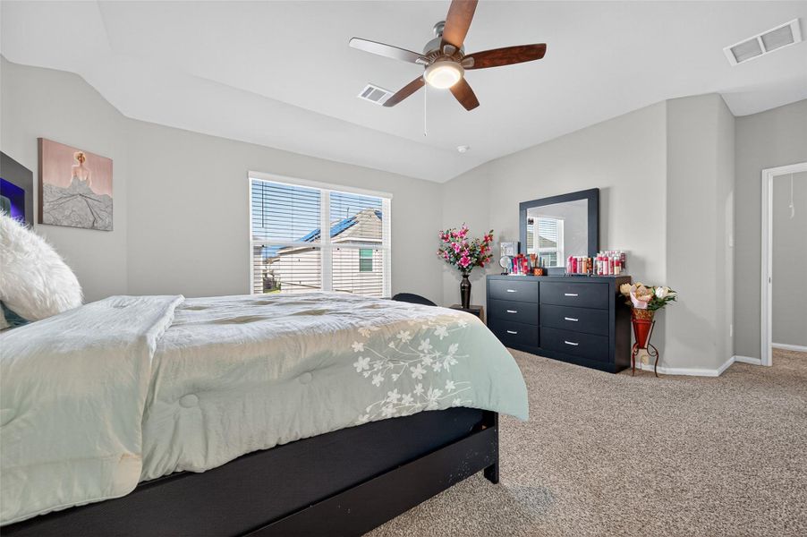 This bedroom is spacious and bright, featuring a large window, neutral walls, and carpeted flooring. It has a ceiling fan, a comfortable bed, and a stylish dresser with a mirror. The room is well-decorated with floral accents, creating a cozy yet elegant atmosphere.