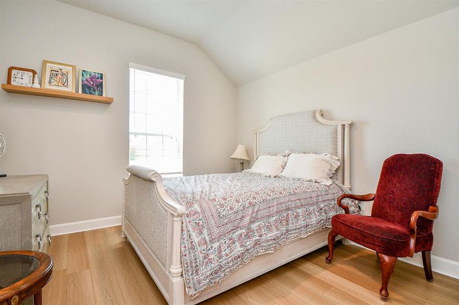 Cozy bedroom with a stylish bed featuring a patterned comforter. The room has a vaulted ceiling, light walls, and wood flooring. A red armchair adds a pop of color, and a window allows natural light to brighten the space.