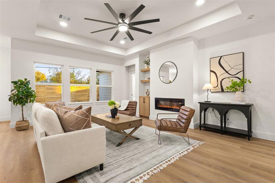 Living area featuring a raised ceiling, a glass covered fireplace, light wood-style flooring, recessed lighting, and ceiling fan
