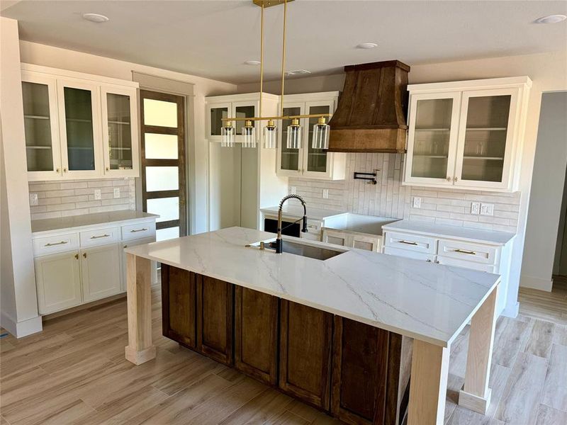 Kitchen with glass insert cabinets, backsplash, premium range hood, light stone counters, and a center island with sink