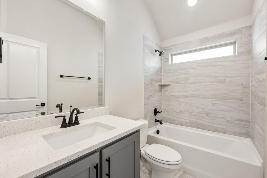 Bathroom with gray vanity, quartz countertop, tub-shower combo with light tile surround, and vaulted ceiling Bathroom with gray vanity, quartz countertop, tub-shower combo with light tile surround, and vaulted ceiling