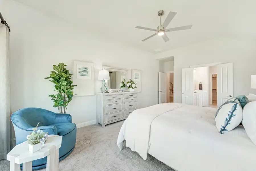 Spacious master bedroom with ceiling fan, white dresser, blue accent chair, and ensuite bathroom access