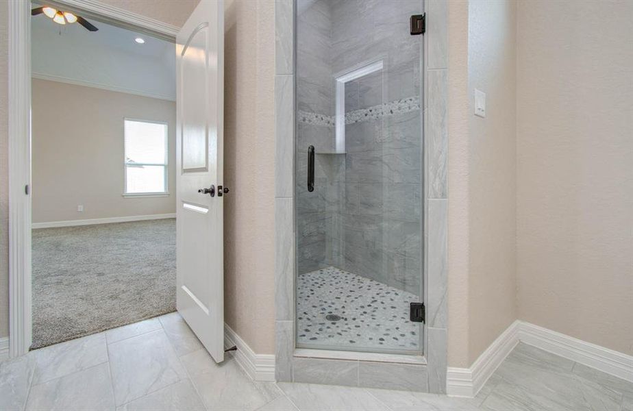 This photo shows a modern bathroom with sleek grey tile work, featuring a spacious walk-in shower with a glass door. The view extends into an adjoining carpeted room, suggesting a seamless flow between spaces.