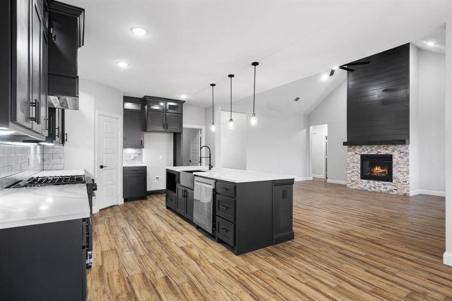 Kitchen with a stone fireplace, backsplash, pendant lighting, vaulted ceiling, and a center island with sink