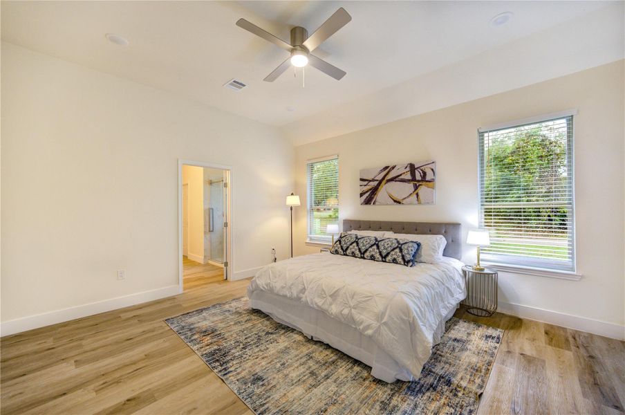 Bright and spacious bedroom with a modern design, featuring a ceiling fan, large windows for natural light, and an ensuite bathroom. The room is accented with wood flooring creating a cozy yet elegant atmosphere.