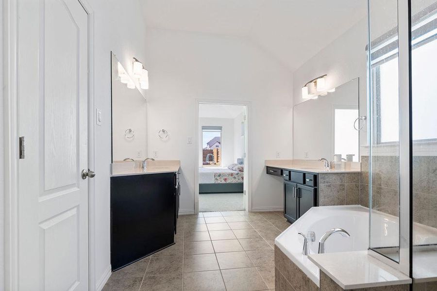 Ensuite bathroom featuring two vanities, a garden tub, lofted ceiling, and light tile patterned floors Ensuite bathroom featuring two vanities, a garden tub, lofted ceiling, and light tile patterned floors