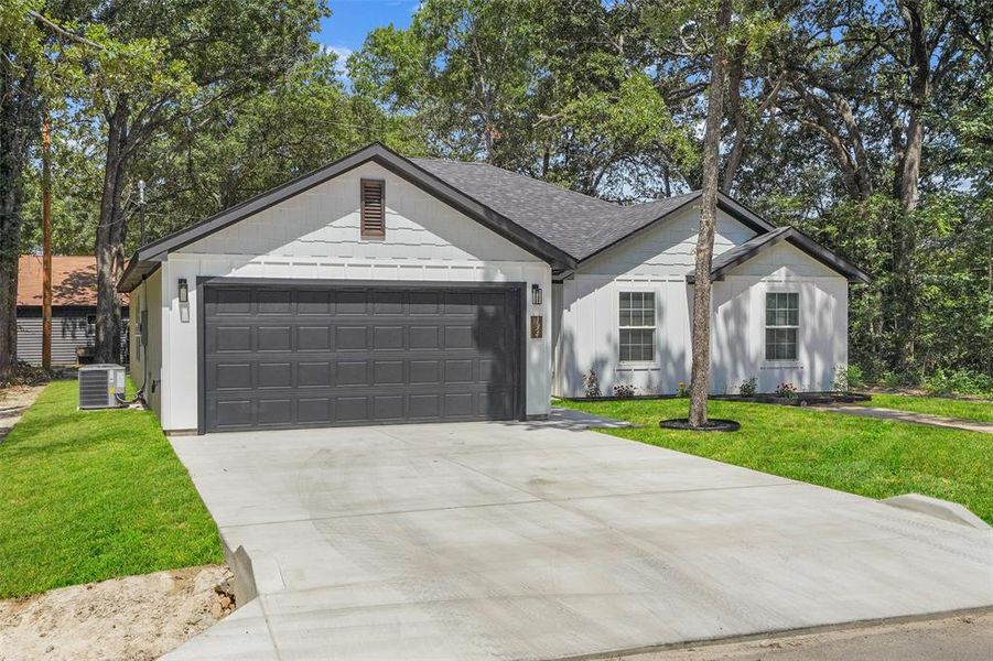 Contemporary single-story residence featuring a dark gray two-car garage door, white horizontal siding, a dark gray shingled roof, a concrete driveway, and established trees Contemporary single-story residence featuring a dark gray two-car garage door, white horizontal siding, a dark gray shingled roof, a concrete driveway, and established trees