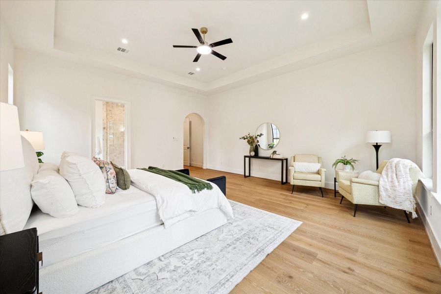 Large primary suite with tray ceiling, white oak wood flooring, sitting area, and access to the ensuite bath.