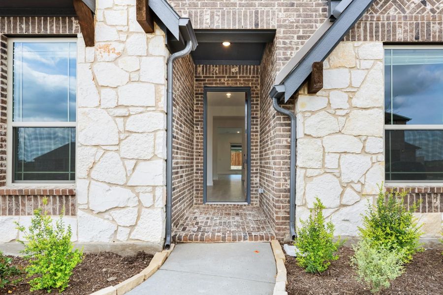 Front entry with brick and stone exterior, covered porch, gray door, and concrete walkway