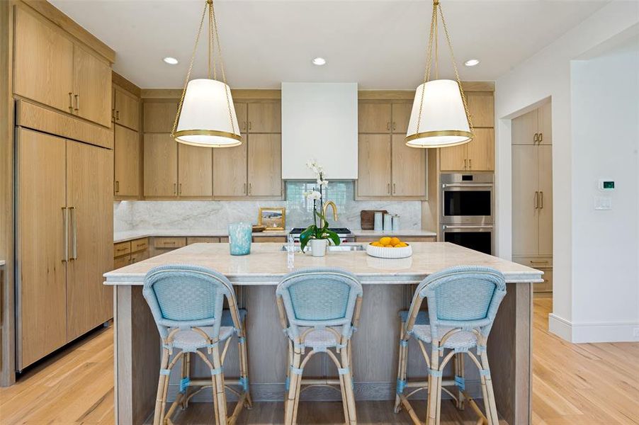 Gourmet kitchen featuring a large central island with a light-toned countertop, integrated panel-front refrigerator, stainless steel double ovens, custom wood-finish cabinetry, and wide plank wood-finish flooring
