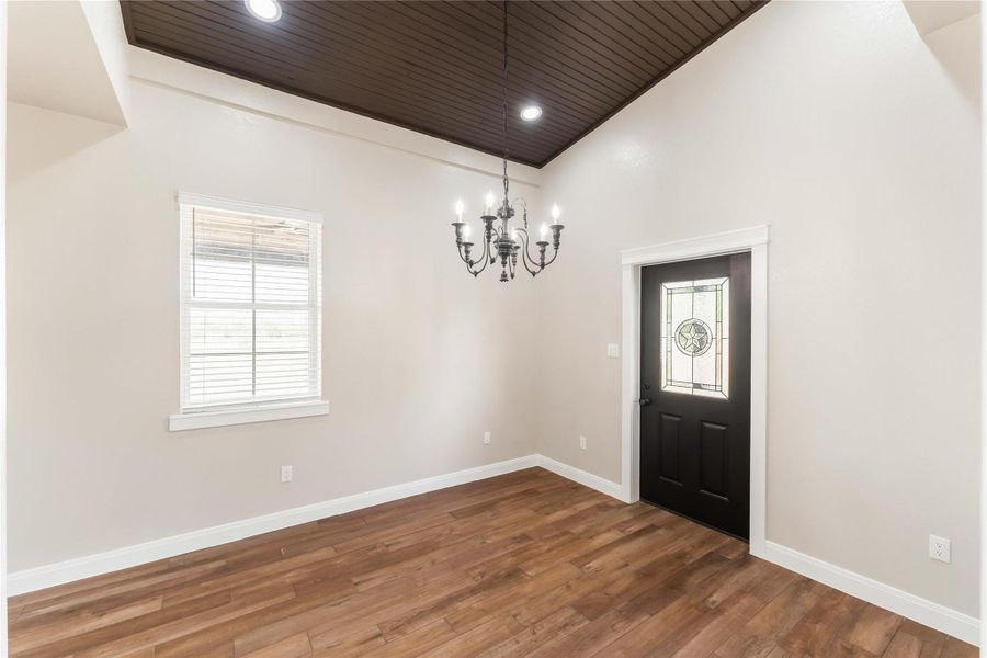 This room features a warm tile wood-style floor and a dark wood ceiling with recessed lighting. It includes a stylish chandelier and a decorative front door with glass detailing, complemented by a window that lets in natural light.