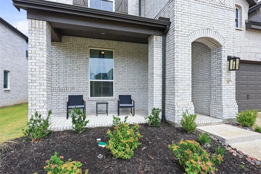 Front facade featuring light gray brick, a covered porch with dark wood-finish columns, an arched entryway, an attached garage with a dark panel door, and dark exterior lighting