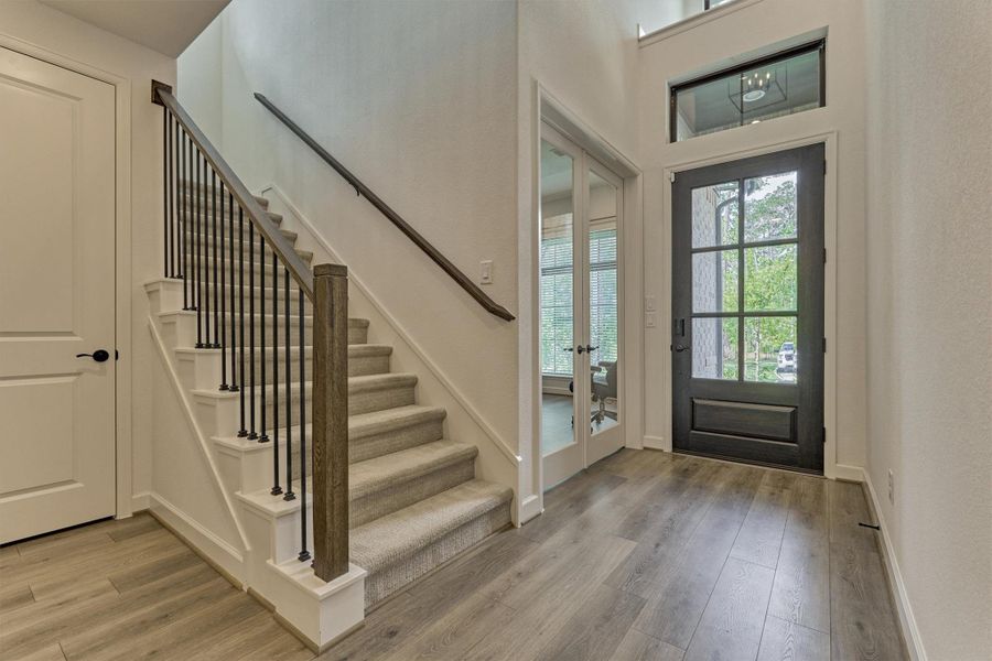 This entryway features a modern design with a dark-framed glass front door, light wood flooring, and a carpeted staircase with sleek black railings. The space is bright and airy, with an adjacent room accessed through double glass doors.