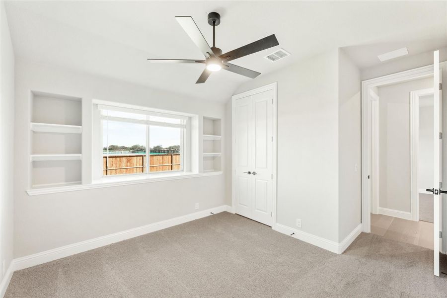 Bedroom with carpet, ceiling fan, built-in shelves, closet, and window overlooking backyard fence Bedroom with carpet, ceiling fan, built-in shelves, closet, and window overlooking backyard fence