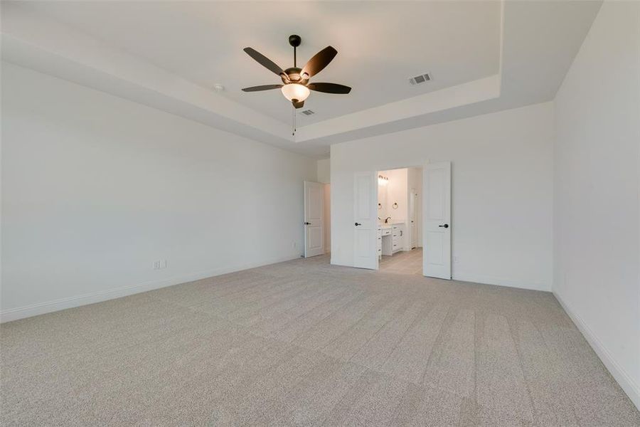 Unfurnished bedroom with a raised ceiling, light colored carpet, ensuite bathroom, and ceiling fan