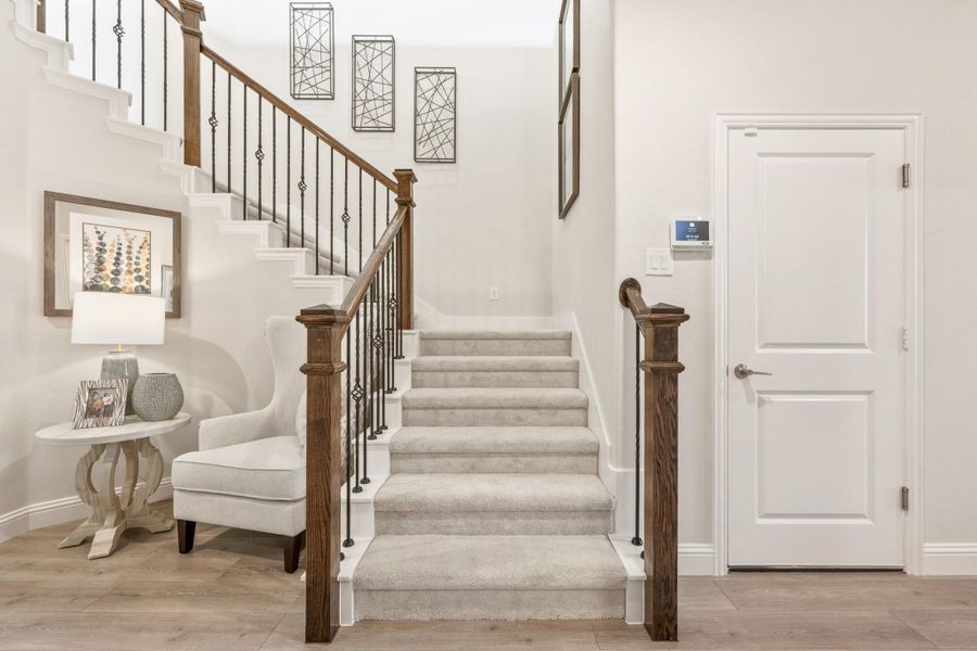 Carpeted staircase with wood newel posts and wrought iron balusters in a bright entryway with accent chair nearby. Carpeted staircase with wood newel posts and wrought iron balusters in a bright entryway with accent chair nearby.