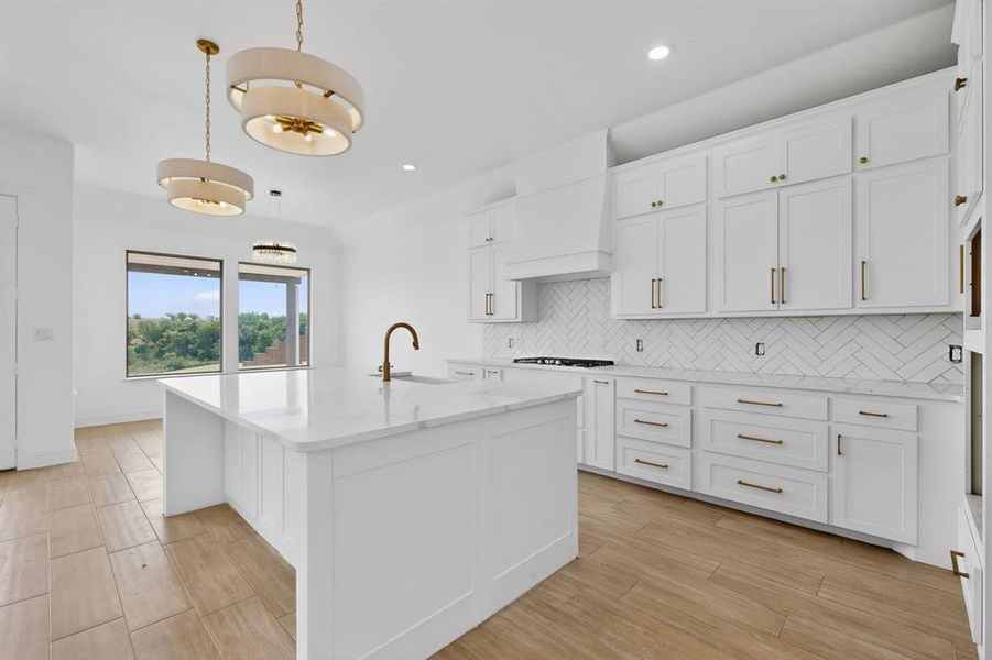Bright kitchen featuring a central island with a built-in sink and brass-finish faucet, white shaker-style cabinetry, and a white herringbone tile backsplash Bright kitchen featuring a central island with a built-in sink and brass-finish faucet, white shaker-style cabinetry, and a white herringbone tile backsplash