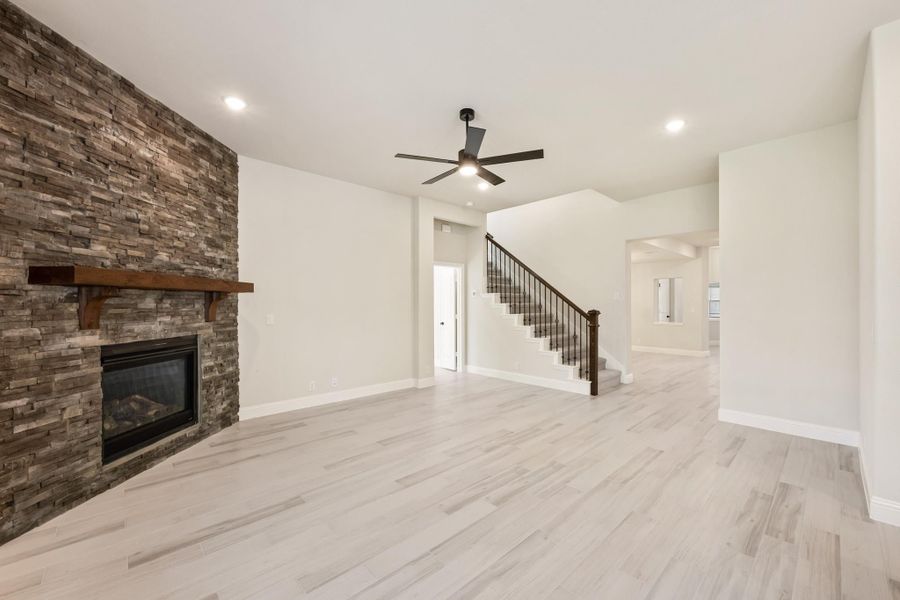 Open living room with stacked stone fireplace, light wood floors, ceiling fan, and staircase with iron railing Open living room with stacked stone fireplace, light wood floors, ceiling fan, and staircase with iron railing