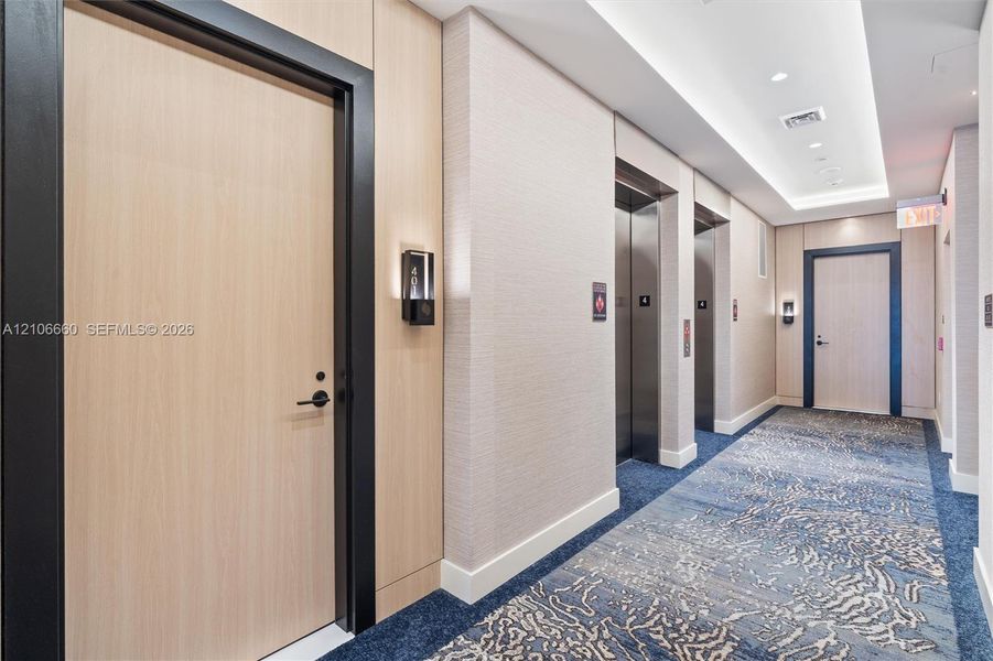 Elevator lobby featuring wood-panel doors with black trim, stainless steel elevator banks, recessed ceiling lighting, and patterned carpeting