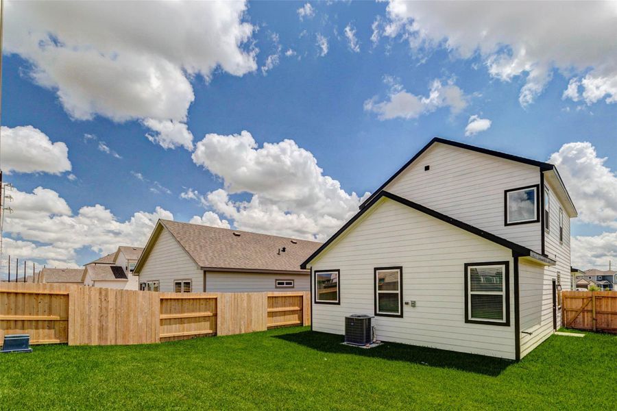 Rear exterior view showcasing the home’s crisp white siding, black trim, fenced backyard, green lawn, and bright sky backdrop. The yard offers usable outdoor space for pets, play, grilling, or relaxing, with side access and a clean new construction exterior.