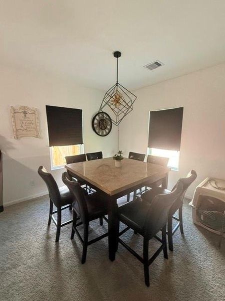 This dining area features a contemporary square table with eight chairs, complemented by a modern geometric light fixture. The room has two windows with shades, neutral walls, and  creating a cozy and stylish setting.