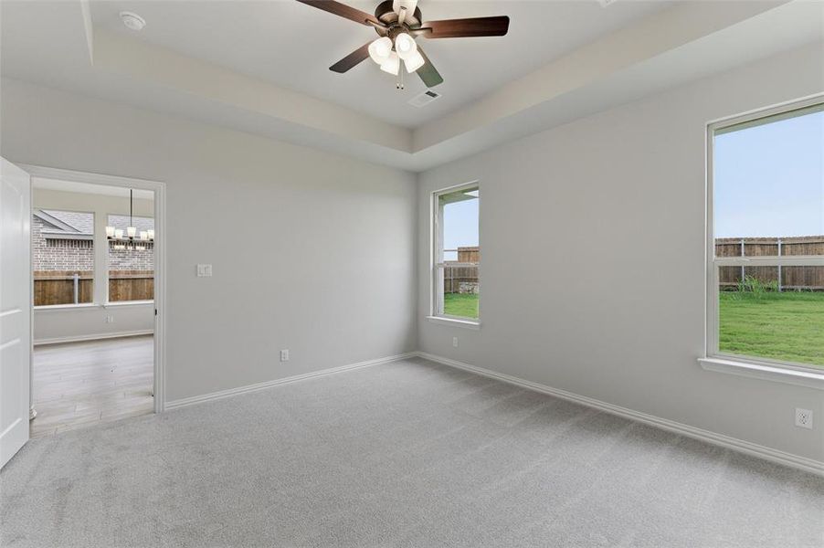 Spacious room featuring light gray carpet flooring, a tray ceiling with a ceiling fan, and two windows offering natural light