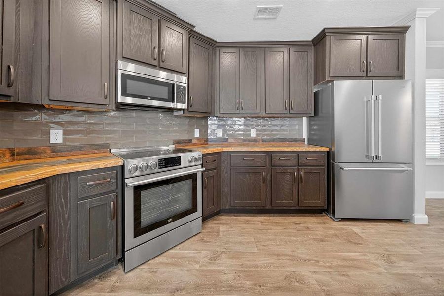 Kitchen with appliances with stainless steel finishes, light wood finished floors, decorative backsplash, butcher block counters, and dark brown cabinets Kitchen with appliances with stainless steel finishes, light wood finished floors, decorative backsplash, butcher block counters, and dark brown cabinets