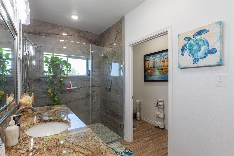 Bathroom featuring a glass-enclosed shower with stone-finish tile surround, a single vanity with a granite countertop and undermount sink, and recessed ceiling lighting Bathroom featuring a glass-enclosed shower with stone-finish tile surround, a single vanity with a granite countertop and undermount sink, and recessed ceiling lighting