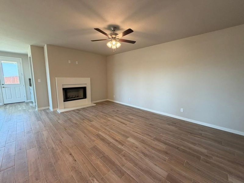 Spacious living area featuring wood-finish flooring, a white mantel fireplace, a ceiling fan with integrated lighting, and a white paneled door with glass insert