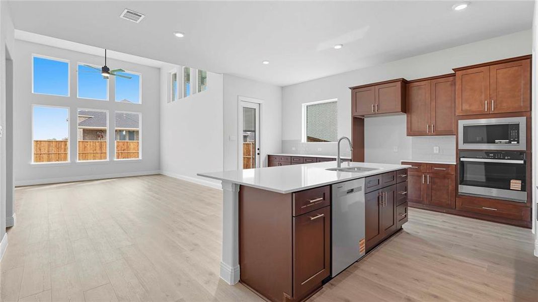 Open-concept kitchen and living area featuring high ceilings, wood-finish flooring, a kitchen island with a built-in sink and dishwasher, dark wood cabinetry, and a suite of stainless steel appliances including a microwave and oven