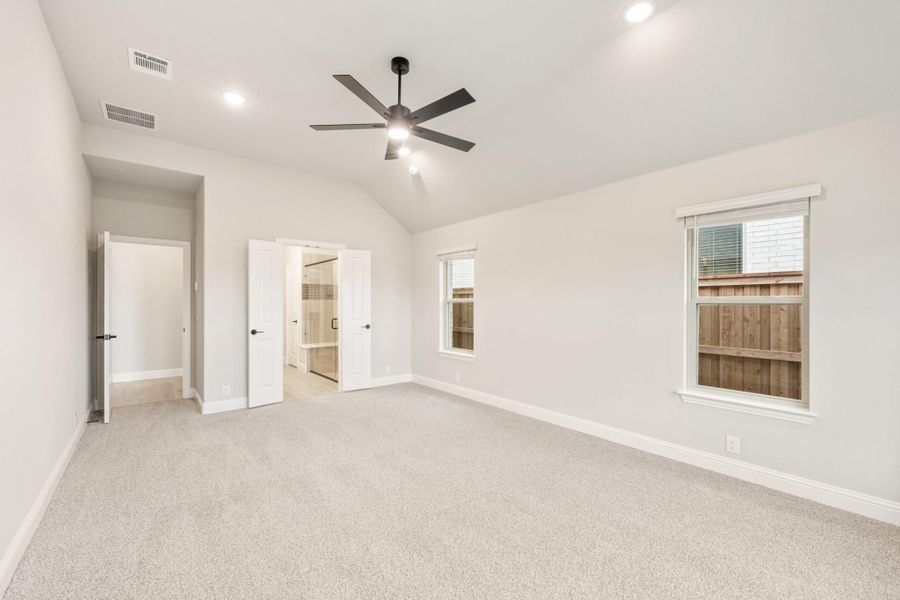 Empty bedroom with vaulted ceiling, ceiling fan, carpet flooring, and ensuite bathroom door open