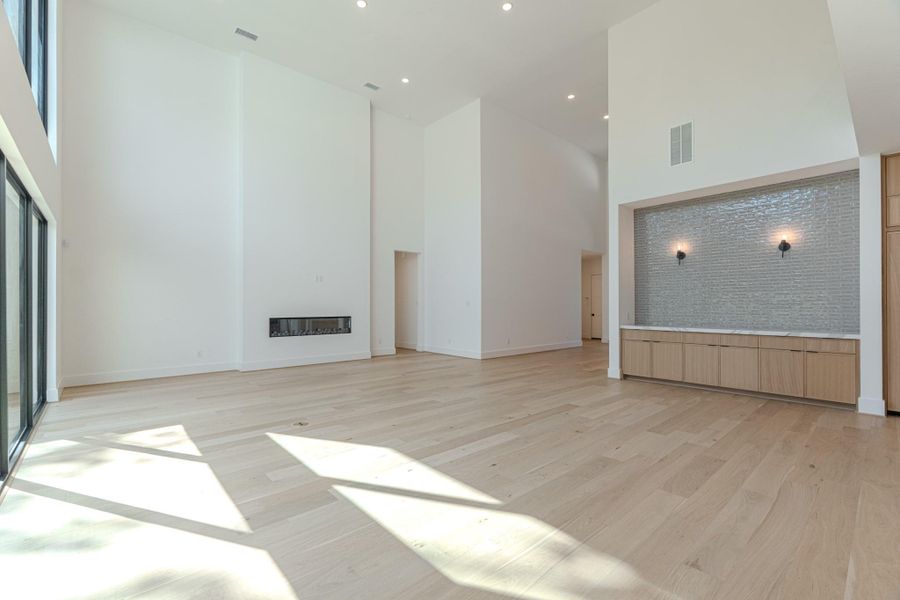 This spacious, modern living area features high ceilings, large windows for abundant natural light, light wood flooring, a sleek built-in fireplace, and a stylish alcove with textured wall and built-in cabinetry.