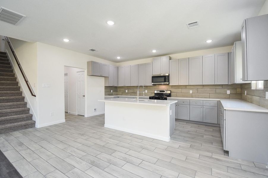Modern kitchen with stainless steel appliances, gray shaker cabinets, and a central island. Open floor plan with wood-look tile flooring extending to the living area. Staircase to the side leading to the upper level. Modern kitchen with stainless steel appliances, gray shaker cabinets, and a central island. Open floor plan with wood-look tile flooring extending to the living area. Staircase to the side leading to the upper level.