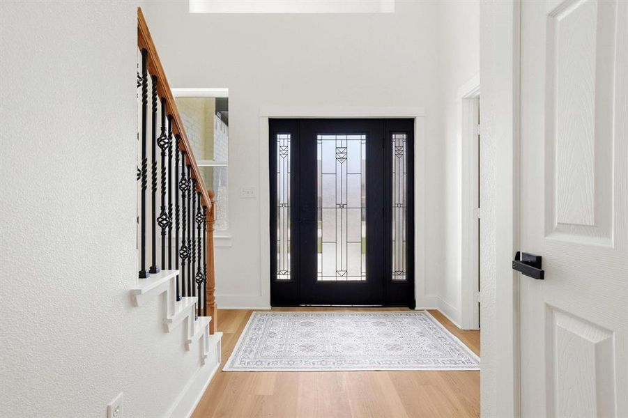 Entryway featuring a decorative transom and sidelight entry door, light wood-finish flooring, white walls, and a staircase with wrought iron balusters and a wood handrail Entryway featuring a decorative transom and sidelight entry door, light wood-finish flooring, white walls, and a staircase with wrought iron balusters and a wood handrail