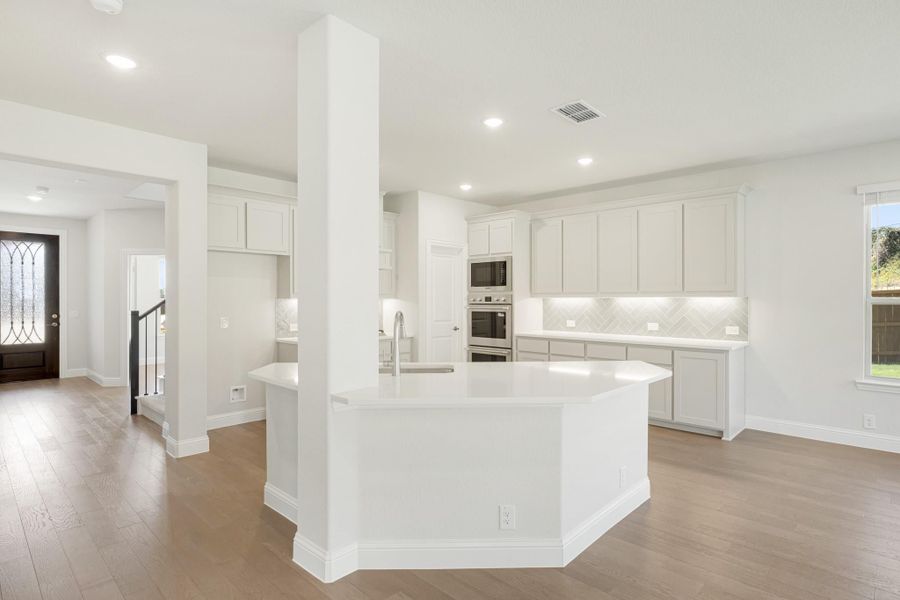 Open kitchen with large white island, gray cabinets, herringbone backsplash, and stainless steel double ovens on wood floors.