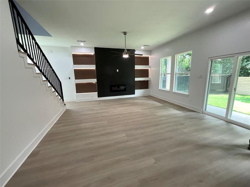 Expansive living area featuring light wood-finish flooring, a modern black fireplace surround with an electric fireplace, and integrated shelving with accent lighting Expansive living area featuring light wood-finish flooring, a modern black fireplace surround with an electric fireplace, and integrated shelving with accent lighting
