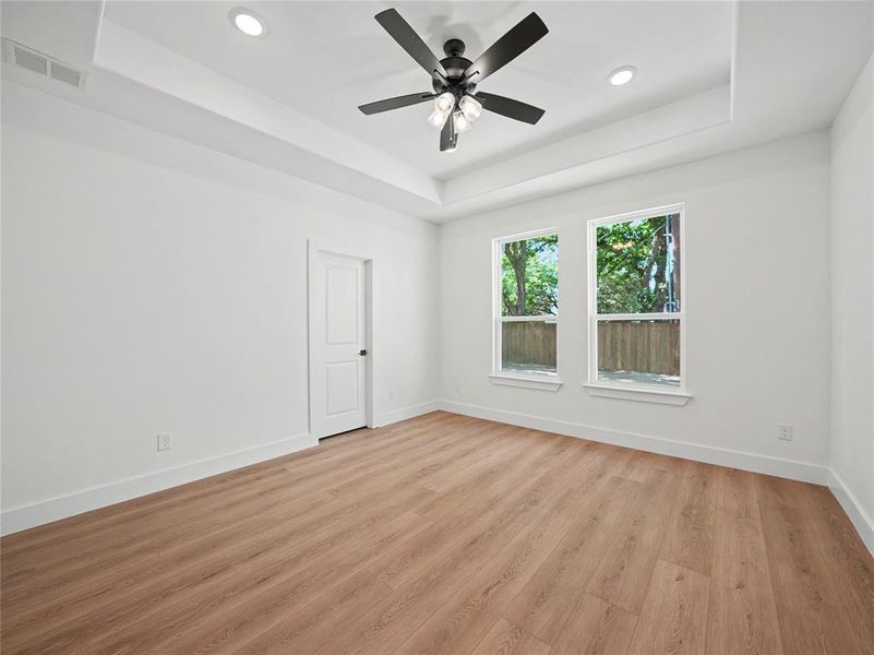 Spacious room featuring wood-finish flooring, two windows, a tray ceiling with recessed lighting, and a ceiling fan with integrated lighting Spacious room featuring wood-finish flooring, two windows, a tray ceiling with recessed lighting, and a ceiling fan with integrated lighting