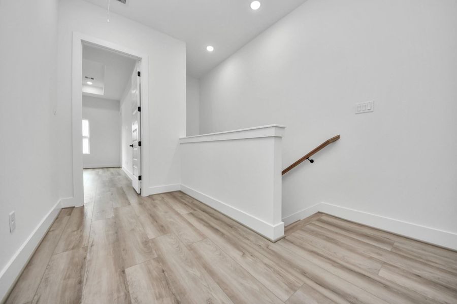 A bright, modern hallway with light wood flooring leads to a room with a white door. There are recessed lights in the ceiling and a simple wooden handrail along the staircase. The space feels open and freshly updated.