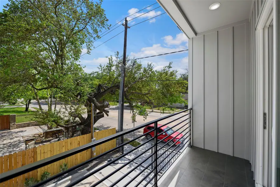 The modern balcony has a sleek metal railing, overlooking a tree-lined street and a wooden fence. The area is bright with natural light, offering a pleasant view of the neighborhood.
