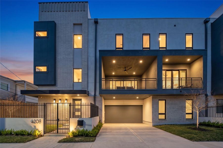 Virtually Enhanced Twilight Image- Illuminated three-story custom home showcasing contemporary brick-and-stucco architecture, a gated entrance, private driveway, and two-car garage. Virtually Enhanced Twilight Image- Illuminated three-story custom home showcasing contemporary brick-and-stucco architecture, a gated entrance, private driveway, and two-car garage.
